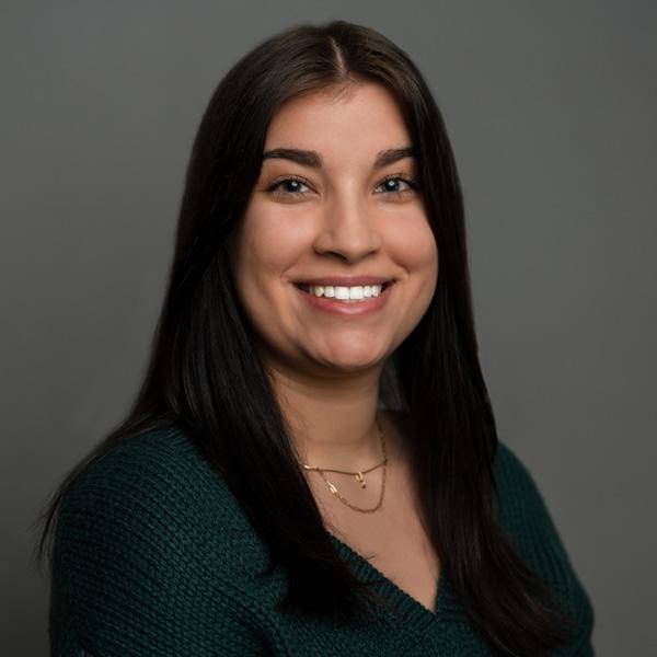 Professional portrait of a smiling woman with long dark hair, wearing a green sweater, against a gray background.